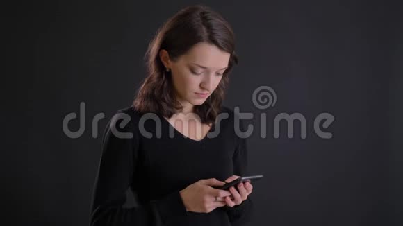 年轻迷人的白种人长发女孩在黑色背景下专心观看智能手机的肖像视频的预览图