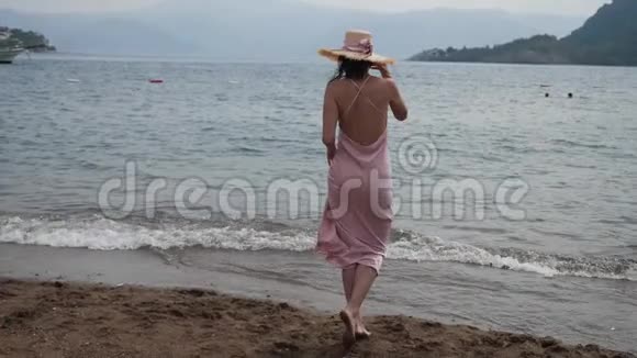 穿着粉红色丝绸裙子和草帽的女人在海滩上散步视频的预览图