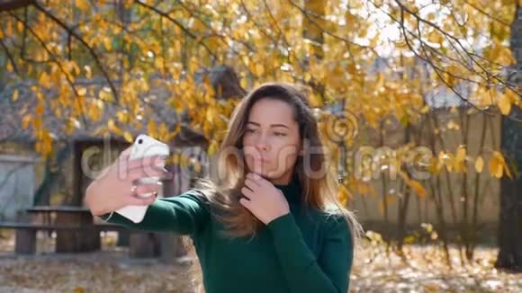 年轻漂亮的黑发女人在秋天的黄树背景下在智能手机上自拍视频的预览图