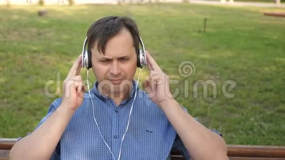 英俊的年轻人带着耳机听着智能手机里的音乐在公园外面的长椅上跳舞视频的预览图
