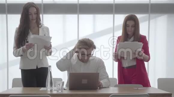 一个戴着眼镜的金发男子坐在一间带上网本的舒适的办公室里两个穿着正式证件的女孩视频的预览图