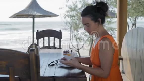 美丽的女人穿着橙色的裙子坐在海边的咖啡馆里在智能手机上留言视频的预览图
