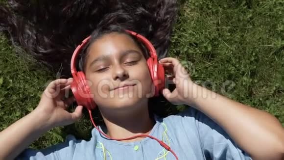美丽的女孩留着长长的黑发躺在公园的草地上听着红色耳机上的音乐视频的预览图
