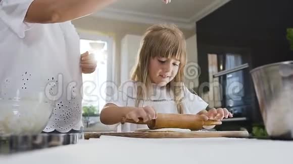 漂亮的妈妈教她年幼的小女儿做饭并在桌子上揉面团做饼干视频的预览图