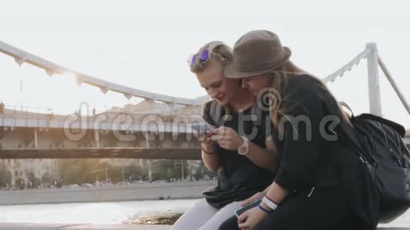 两个年轻的少女坐在河堤上用手机视频的预览图