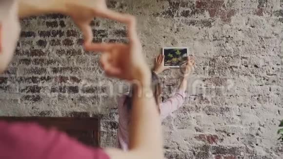 漂亮的亚洲女人在砖墙上选择相框照片的位置而她的男朋友则在做相框造型视频的预览图
