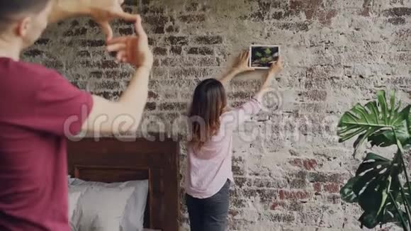 有魅力的一个女孩在砖墙上选择拍照的位置而她的丈夫正在用他的画框造型视频的预览图