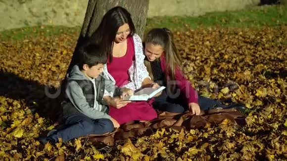妈妈和孩子们坐在树下看书秋天森林里的妈妈和孩子们他们很开心视频的预览图