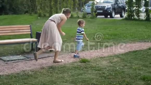 在公园里穿裙子的年轻母亲跑着追着她那爱笑的蹒跚学步的男孩视频的预览图