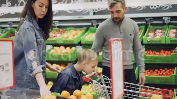 年轻的家庭有着快乐的孩子带着购物车走在食品店里选择水果闻着气味视频的预览图