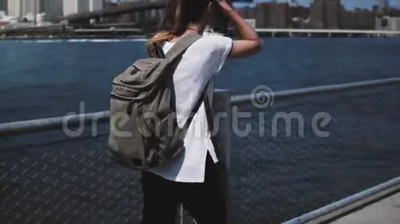 镜头跟随年轻的女游客背着背包轻松地走向阳光明媚的河岸上的布鲁克林大桥视频的预览图