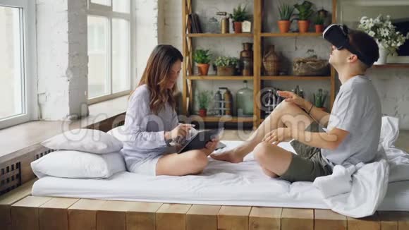 年轻夫妇正在玩男人戴着虚拟现实眼镜而女人正在触摸平板电脑屏幕他们是视频的预览图