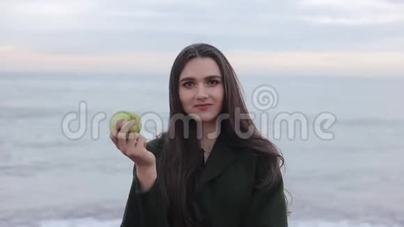 一个美丽的女孩在海滩上吃新鲜苹果的近景一个穿绿色外套的女孩在海边视频的预览图