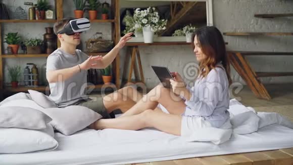 年轻男子使用虚拟现实眼镜而他迷人的妻子正在触摸平板电脑屏幕看着她的丈夫视频的预览图