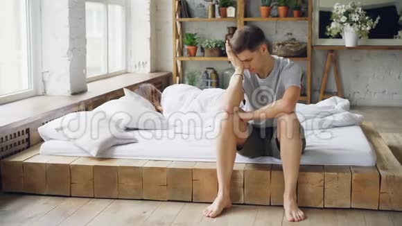 一个悲伤的男人坐在床上和妻子吵了一架摸着他的脸看着窗户而他的妻子躺在床上视频的预览图