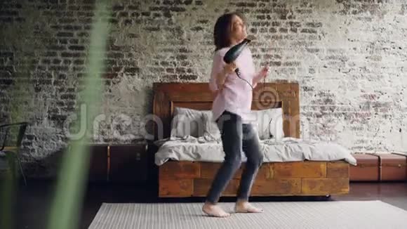 有趣的女孩正在跳舞和唱歌拿着吹风机在卧室的地毯上玩得很开心靠近双人床活跃的年轻妇女视频的预览图