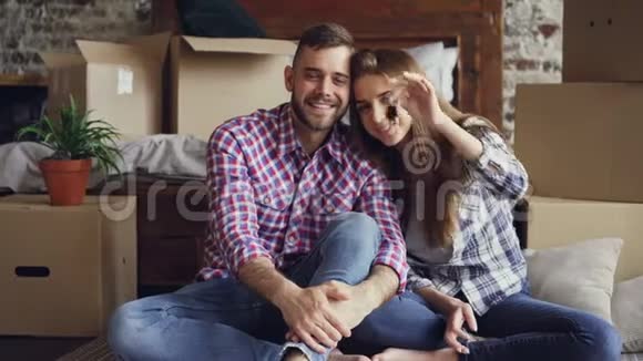 幸福的年轻夫妇坐在新公寓里拿着钥匙看着相机的肖像不错的阁楼风格视频的预览图