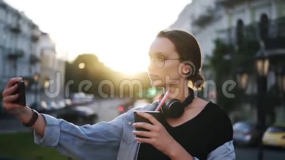 一幅年轻迷人的女人戴着太阳镜的肖像脖子上戴着耳机摆在城市街道上的摄像机上关闭视频的预览图