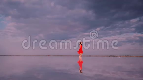 年轻女子穿着一件挥动的连衣裙沿着一个粉红色的湖走湖中的云彩被反射出来视频的预览图