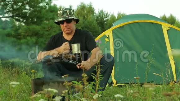 一个人坐在帐篷旁喝茶把肉放在烤架上视频的预览图