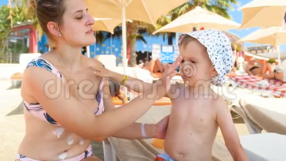 4k镜头微笑的母亲保护她的孩子免受阳光紫外线的照射并在海滩上涂上防晒乳液视频的预览图