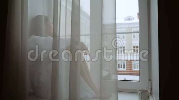 一个穿着白色外套的沮丧的女人坐在窗台上向外看着街道外面正在下雪一片悲伤视频的预览图