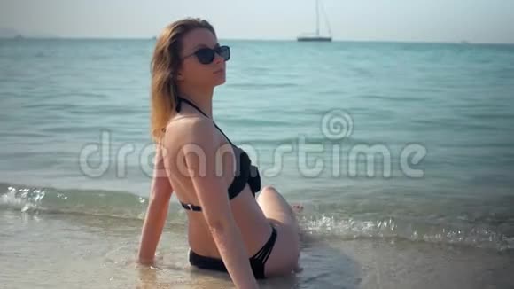 年轻女孩穿着黑色比基尼在阳光明媚的日子里在海滩上放松后景坐在水里视频的预览图
