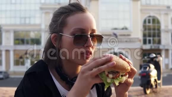 年轻的女性用两只手吃一个大而多汁的汉堡包有短发和深色太阳镜的年轻女孩很饿关闭视频的预览图