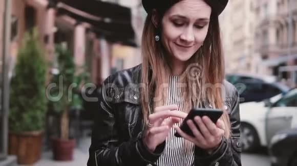 微笑快乐的白种人女性博主戴着时尚的帽子沿着街道走使用智能手机购物应用程序慢动作视频的预览图