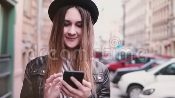 微笑的年轻欧洲妇女戴着时尚的帽子走在城市的街道上带着智能手机期待着缓慢的运动视频的预览图