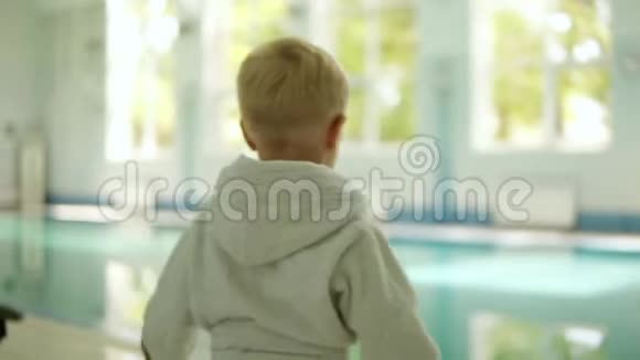 一个穿着浴袍的小男孩从两只手中把球扔进游泳池的背面很好玩在外面视频的预览图