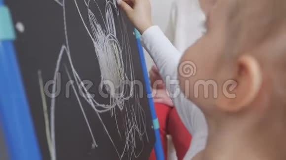 小女孩用粉笔在托儿所的黑板上画画妈妈坐在她旁边佩服这个孩子视频的预览图