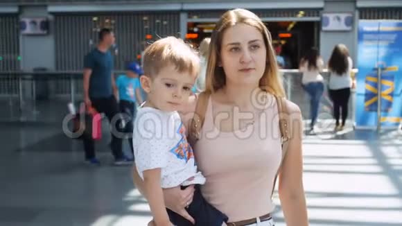 慢动作视频年轻微笑的女人抱着她蹒跚学步的儿子在现代机场航站楼散步视频的预览图
