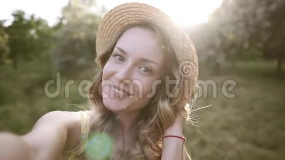 美丽的金发女人戴着草帽自拍摆姿势拍照送空气吻背景上阳光明媚的草地视频的预览图