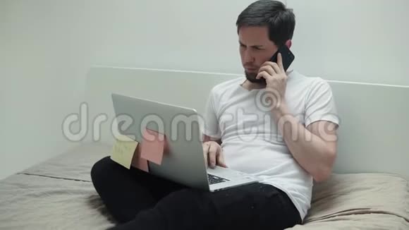 一个忙碌的男人坐在卧室里用手机打着笔记本视频的预览图