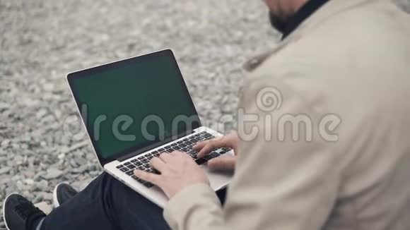 男人坐在沙砾滩上拿着彩色键盘的笔记本工作视频的预览图
