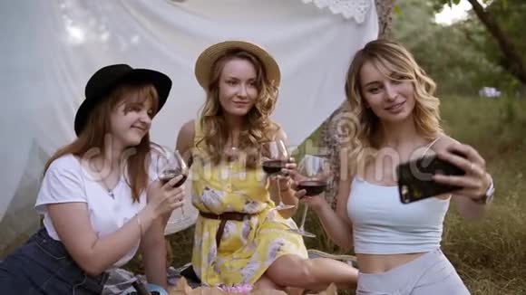 三个漂亮的现代女人坐在草地上端着一杯红酒一个金发女孩和她一起自拍视频的预览图