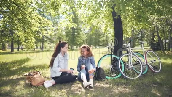 迷人的年轻女性游客正在公园里喝咖啡坐在草地上社交他们的自行车和背包视频的预览图