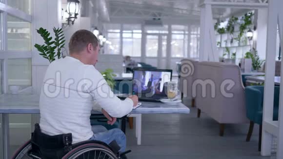 坐在轮椅上的学生残疾人在笔记本上写笔记同时坐在桌子旁观看在线培训视频的预览图