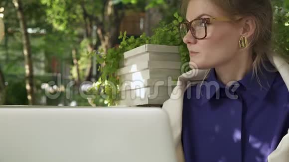 戴眼镜的嬉皮士女孩在她的笔记本电脑附近的夏日咖啡馆喝咖啡视频的预览图