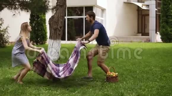 公园里的一对年轻夫妇结束了野餐折叠了一个格子在草地上有一个柳条篮子和水果绿色公园树木视频的预览图