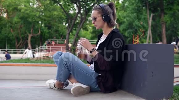 时尚年轻的女孩坐在公园的地板上她的腿交叉喝了一杯软饮料一个戴着太阳镜的女人视频的预览图