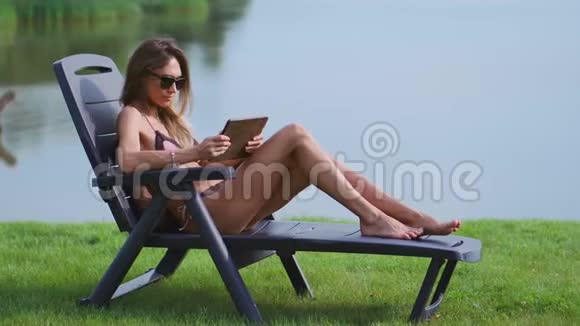 苗条的女人戴着太阳镜躺在一个穿着泳衣的阳光休息室里用平板电脑手指触摸屏幕视频的预览图