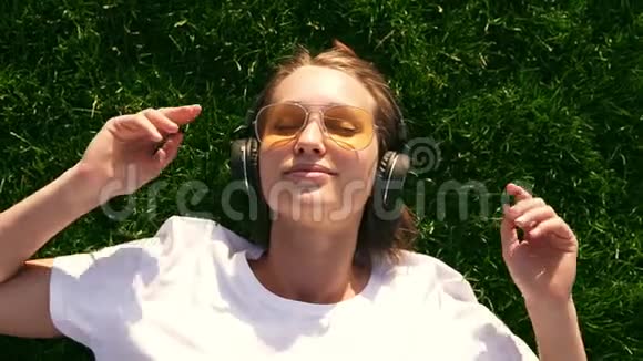 夏天女孩在草地上用耳机听音乐绿草如茵戴着黄色太阳镜的微笑女孩视频的预览图