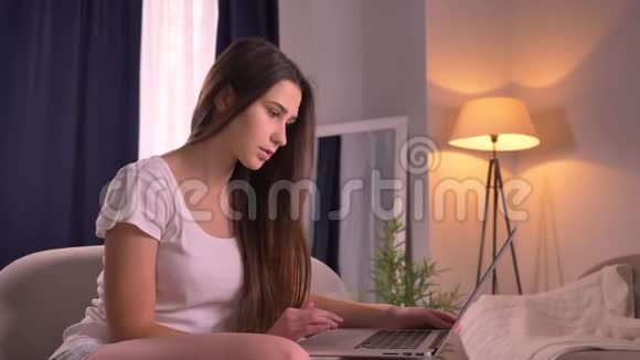 年轻美丽的白种人少女坐在舒适的室内沙发上浏览笔记本电脑的特写照片视频的预览图