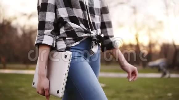 苗条而温柔的女孩穿着牛仔裤和衬衫走在外面绿色的一边右手拿着笔记本电脑光明的一天是视频的预览图