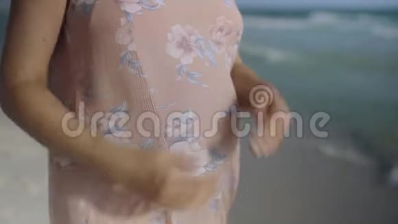 年轻的孕妇站在蓝色海洋附近的海滩上呼吸靠近手和腹部暑假假期视频的预览图