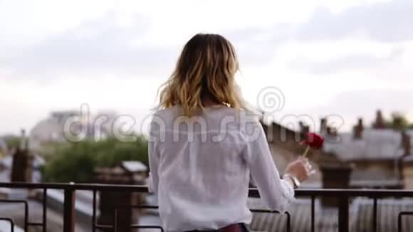 穿着牛仔裤和白衬衫的性感苗条的女人站在阳台上手里拿着红花金发女孩视频的预览图