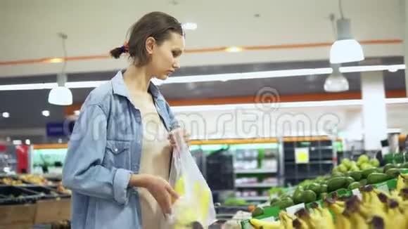 在超市挑选水果时穿衬衫的漂亮女孩的手持镜头把一些香蕉放进玻璃纸袋视频的预览图