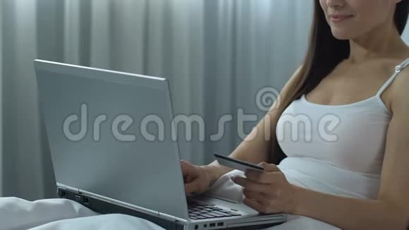 年轻女子在笔记本电脑上输入信用卡号码坐在床上在网上购物视频的预览图